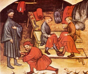 La imagen muestra cuatro personas vestidas con ropas antiguas. Dos de ellas están cosiendo.. En la habitación se ven rollos de tela y tijeras sobre una mesa. 